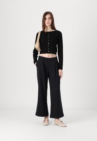 Μαύρη ριμπ κοντή καρντάρκα με κουμπιά μπροστά, συνδυασμένη με φαρδιά σκούρα παντελόνια και ανοιχτόχρωμα επίπεδα παπούτσια. Απλή μπεζ τσάντα χιαστί.