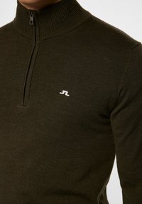 Khaaki vetokirjakkaita, jossa on ribattu kaulus ja hihansuut, sekä pieni valkoinen logo rinnassa. Valmistettu hienosta neuloskankaasta.