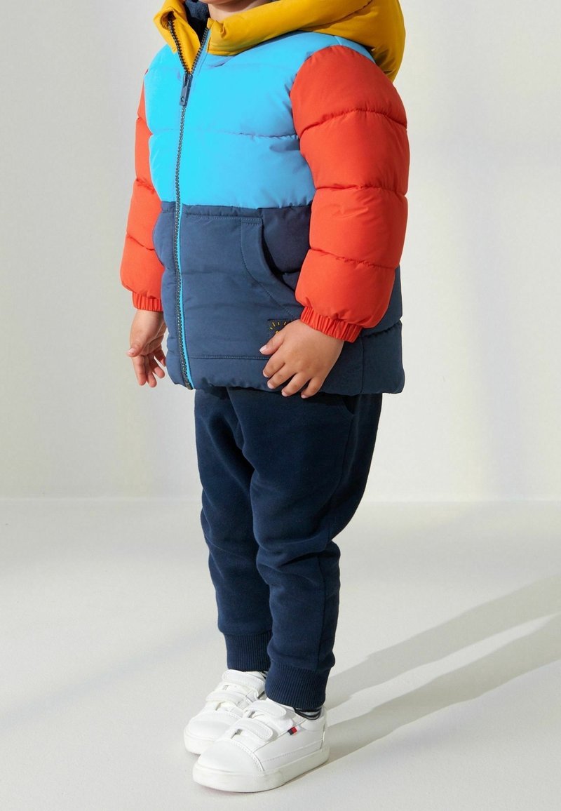 Giacca a vento colorata con sezioni blu, arancioni e gialle. Pantaloni scuri e sneakers bianche. Sfondo chiaro con ombre morbide.