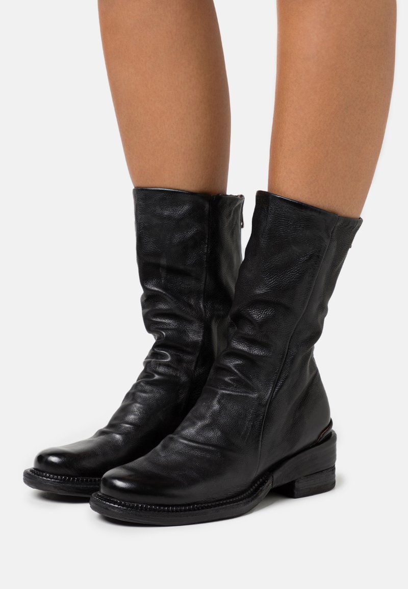 Bottes en cuir noir mi-mollet avec une texture légèrement froissée, portées sur jambes nues, avec un petit talon et un bout arrondi.