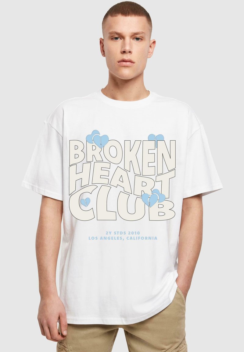 Fehér pamut póló, amelyen nagy méretű grafikai szöveg található: "BROKEN HEART CLUB" világosszürkében, kék szív motívumokkal. Laza szabású, rövid ujjú.