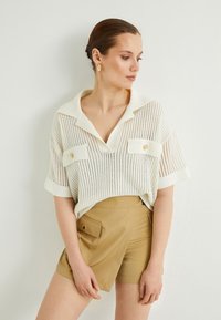 Vit stickad skjorta med krage och två framfickor, tillsammans med beige shorts med hög midja och knappdetalj.