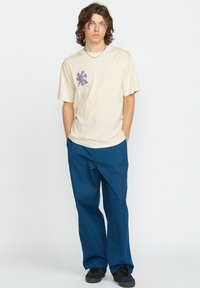 Camisa de color crema con un diseño geométrico azul y naranja en el pecho, combinada con pantalones anchos de color azul oscuro y zapatillas negras.