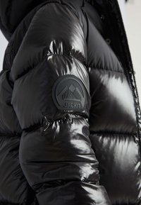 Chaqueta de plumas negra con un acabado brillante, textura acolchada y un parche negro redondo con un diseño de montaña en la manga izquierda.