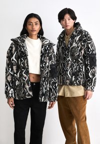 Faux fur jackets with a black, white, and taupe abstract pattern; featuring a high collar and zippered pockets on the sleeves.