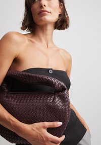 Vävd läderhandväska i djup burgundy med en mjuk textur. Har ett distinkt, böjt handtag och dragkedjestängning.