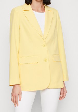 Femme portant un blazer cintré jaune pâle avec deux boutons et des poches à rabat sur un top blanc et un pantalon blanc, debout devant un fond uni.