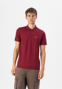 Burgundské polokošile s krátkým rukávem a límečkem, s knoflíkovou légou na tři knoflíky a malým vyšitým logem na hrudi.