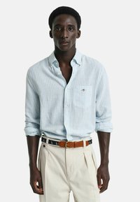 Camicia azzurra a righe con taschino, maniche arrotolate, abbinata a pantaloni color crema e una cintura marrone con motivi di barche a vela.