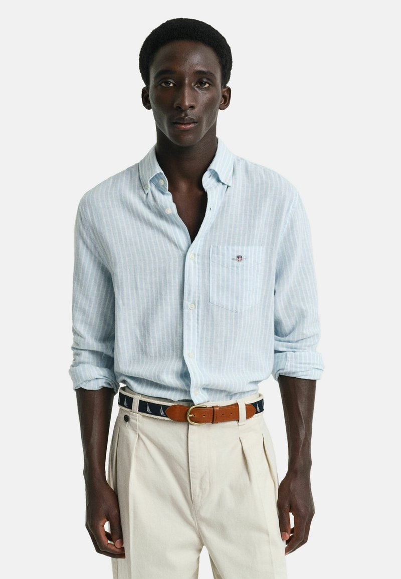 Camicia azzurra a righe con taschino, maniche arrotolate, abbinata a pantaloni color crema e una cintura marrone con motivi di barche a vela.