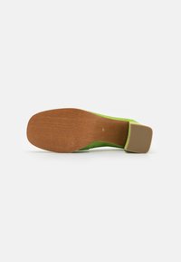 Chaussure à talon en daim vert avec un talon carré et une semelle marron texturée. L'intérieur présente une doublure lisse avec des détails de couture minimalistes.