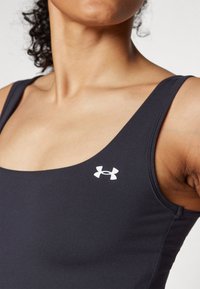 Črn športni top z izrezom in tankimi naramnicami, z belim logotipom Under Armour na levi strani. Gladka tekstura.