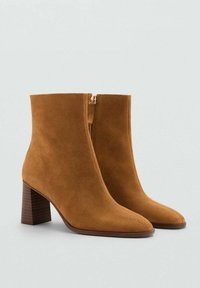 Stivaletti alla caviglia in suede beige con chiusura a zip, punta affusolata e tacco in legno a strisce, spesso. Texture liscia con un design minimalista.