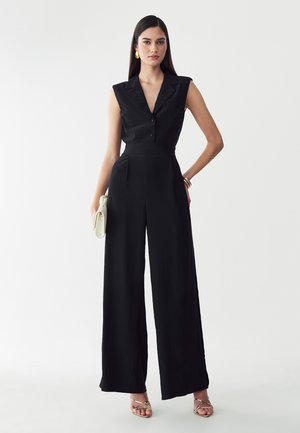 Frau mit langen dunklen Haaren trägt einen ärmellosen schwarzen Jumpsuit mit weitem Bein, goldene Sandalen, goldene Ohrringe, Armbänder und hält eine weiße Clutch.