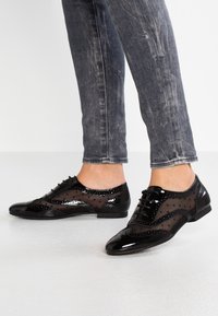 Personne portant des chaussures oxford en cuir laqué noir et en mesh avec des pois noirs, ainsi que des jeans slim gris délavé, se tenant sur un sol blanc.
