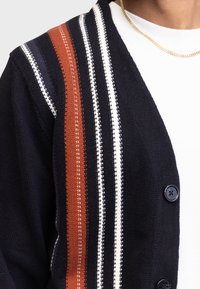 Cardigan bleumarin cu accente în dungi portocalii, albe și roșii, având un material tricotat texturat și nasturi negri.