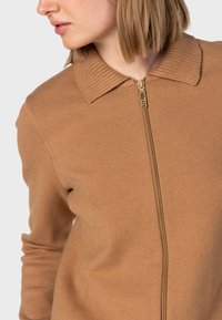 Brun zip-up sweater med ribbet krave, lavet af blødt materiale. Har en frontlynlås, sømløst design og tætsiddende lange ærmer.