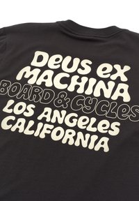 Black t-shirt featuring large white text: “DEUS ex MACHINA BOARD & CYCLES LOS ANGELES CALIFORNIA.” Soft cotton material, crew neck design.