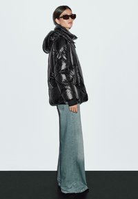 Giubbotto imbottito nero con cappuccio, materiale lucido, vestibilità oversize, tasche laterali, abbinato a jeans svasati azzurri chiari e occhiali da sole scuri.