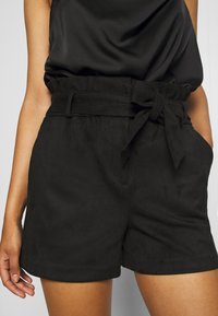 Vrouw in hooggezette zwarte shorts met een gestrikt riempje en zakken, gecombineerd met een zwart mouwloos topje.