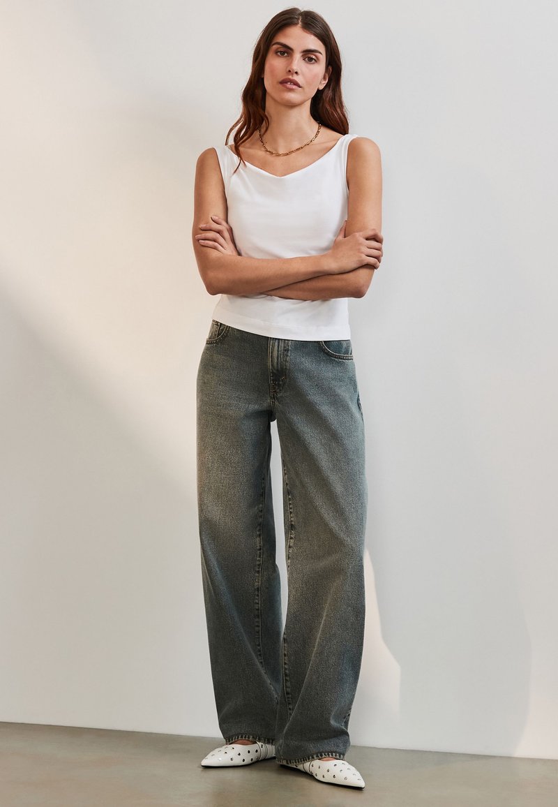 Haut blanc sans manches avec un décolleté en V, associé à un jean en denim gris ample. Chaussures blanches à bout pointu avec des trous.