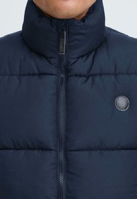 Jūras zila pufīga veste ar augstu apkakli un priekšējo rāvējslēdzēju. Kreisajā pusē ir apaļa logo nozīme. Materiāls izskatās gluds un mīksts.