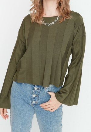 Kvinde iført olivengrøn ribstrikket sweater med viddeærmer og kædehalskæde, kombineret med højttaljede blå jeans med fire knapper.