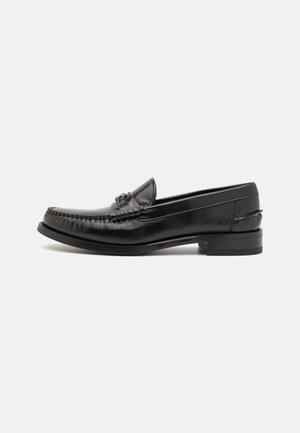 Schwarzer Leder-Slipper mit runder Zehenform, dekorativer Naht und einem kleinen Metallelement auf dem Riemen. Flache Gummisohle.