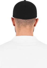 Fekete sapka hátulról nézve, sima textúrával és kerek koronával, egy fehér póló fölött viselve, ami bordázott gallérral rendelkezik.
