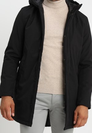 Mand iført en sort hættetrøje over en beige rullekravesweater og lysegrå bukser, stående med armene afslappet ned langs siderne.