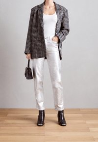 Blazer de patrón blanco y negro sobre una camiseta blanca, combinado con pantalones plateados brillantes y botas de tobillo negras, sosteniendo un pequeño bolso negro.