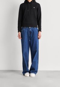 Felpa nera con tasca a marsupio, abbinata a jeans di denim blu larghi. Sneakers bianche completano l'outfit, caratterizzato da una vestibilità rilassata e uno stile informale.