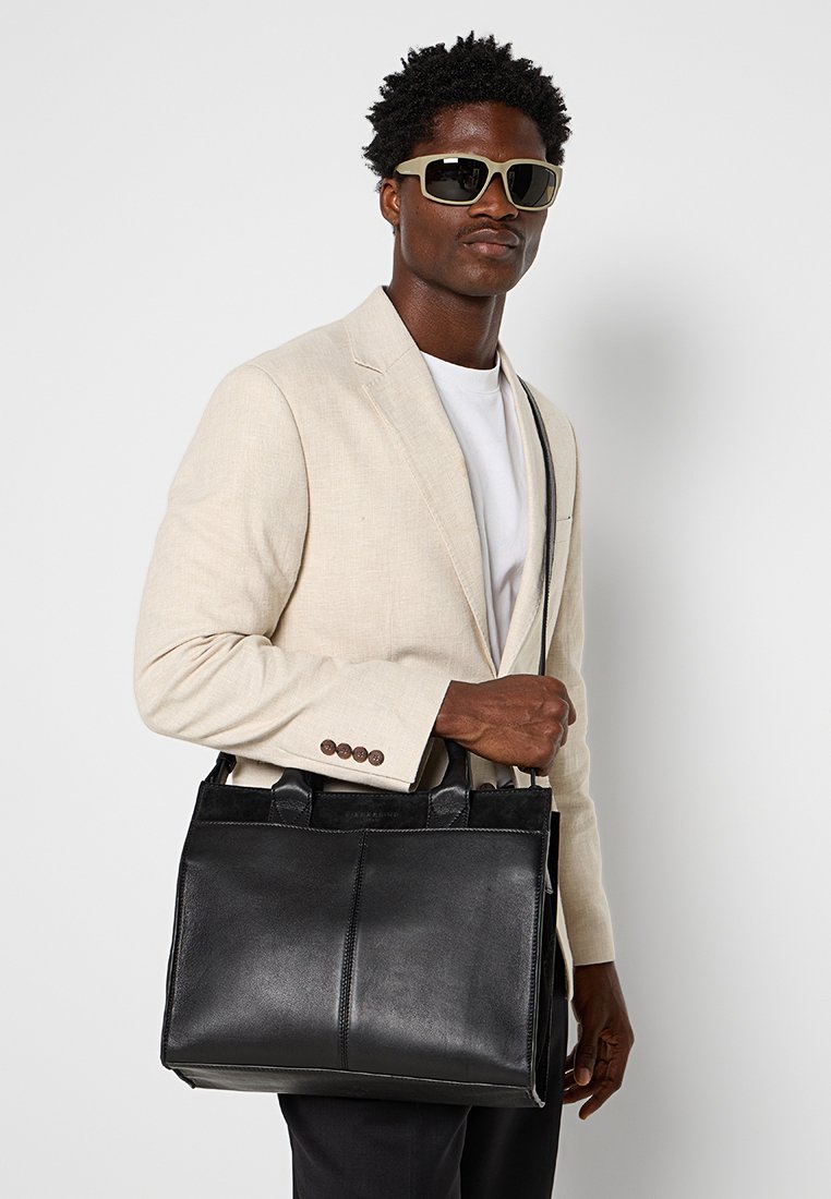 Schwarze Leder-Tote mit doppelten Griffen, rechteckiger Form und Kontrastnähten. Das Modell trägt einen hellen Blazer und Sonnenbrillen.