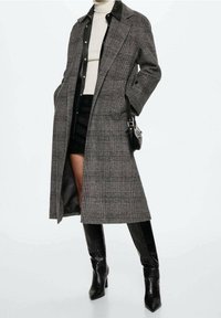 Manteau long à carreaux gris avec un revers noir, présentant un design droit. Porté sur un haut blanc et une jupe noire, accompagné de bottes noires montantes jusqu'au genou.