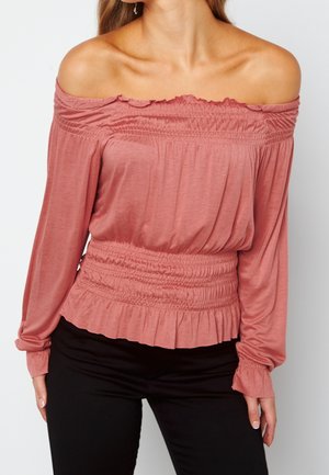 Mujer vistiendo una blusa rosa polvo con hombros descubiertos y fruncida, de mangas largas, combinada con pantalones negros sobre un fondo liso y claro.