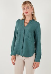 Žalia ilga rankovė bluzė su švelnia tekstūra, turinti dvi priekinės kišenes, užsegimą mygtuku ir apvalų apvadą.