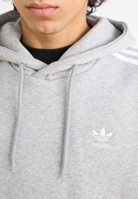 Pelēks kapuce ar teksturētu apdari, ar saņemamu auklu un trim baltām strīpām uz pleciem. Uz krūtīm ir neliels balts "adidas" logo.