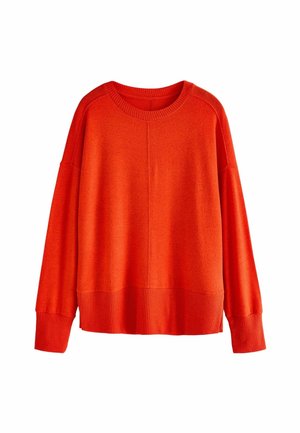 Personne portant un pull orange vif à manches longues rentré dans un jean noir à motifs avec une ceinture noire et une boucle argentée, main dans la poche.
