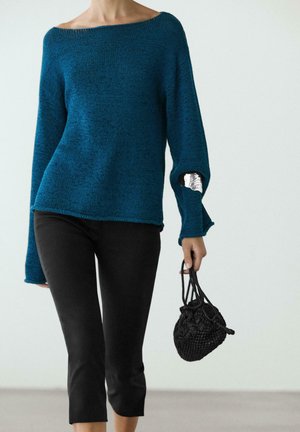 Personne portant un pull bleu sarcelle à manches longues avec un patch argenté sur la manche et un pantalon court noir, tenant un petit sac noir tissé.