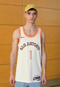 Camiseta de baloncesto blanca con acentos en naranja y negro, que presenta el texto "SAN ANTONIO" y el número 1. Combinada con una gorra amarilla clara.