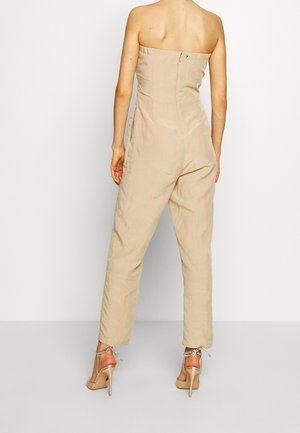 Mono beige sin tirantes con rayas verticales rosas, corpiño ajustado, pantalones ajustados y detalle de cremallera trasera. Combinado con sandalias de tacón altas color beige.