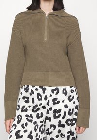 Khakifarbener, gerippter Pullover mit halbem Reißverschluss am Kragen, langen Ärmeln und strukturiertem Strickmuster. Kombiniert mit weißen Hosen mit schwarzem Leopardenmuster.