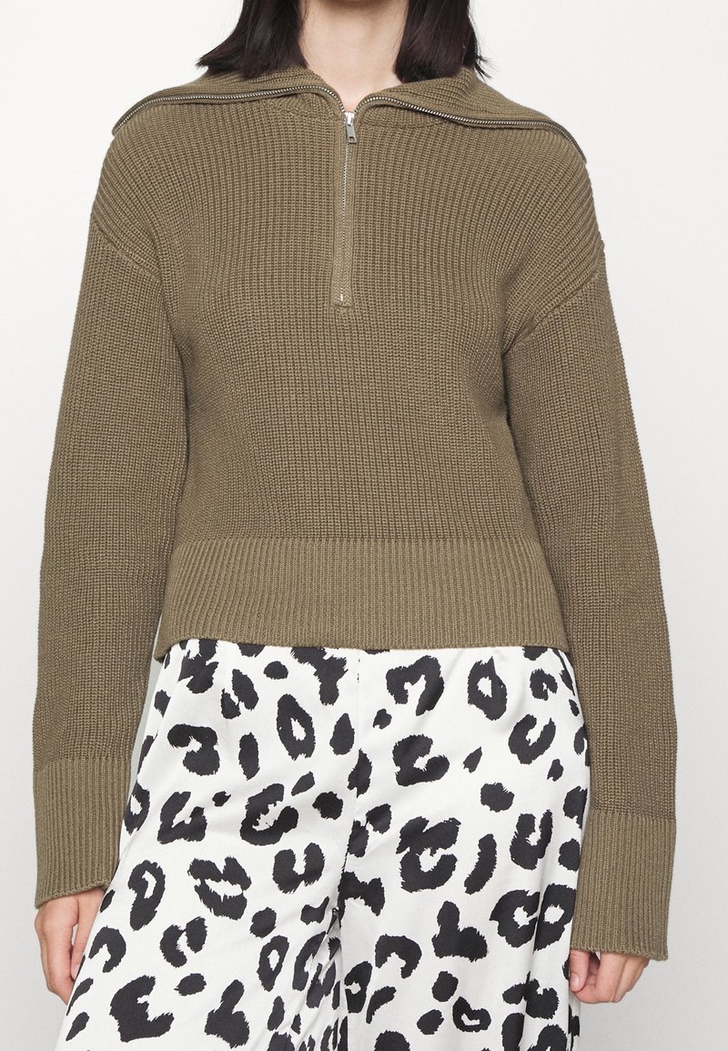 Khakifarbener, gerippter Pullover mit halbem Reißverschluss am Kragen, langen Ärmeln und strukturiertem Strickmuster. Kombiniert mit weißen Hosen mit schwarzem Leopardenmuster.