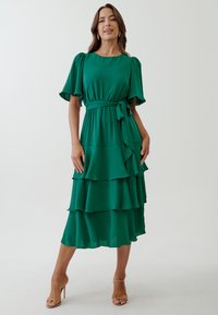 Green tiered dress with short flutter sleeves, a waist belt, and a flowing silhouette. Made of lightweight fabric with a soft texture.