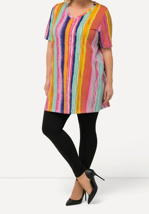 Mujer con una túnica de rayas coloridas con mangas cortas, leggings negros y zapatos de tacón negro, de pie con las piernas cruzadas.