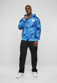 Hoodie bleu tie-dye avec une fermeture éclair à l'avant, des manches élastiques et un détail logo blanc. Associé à un pantalon cargo noir ample et des baskets blanches.