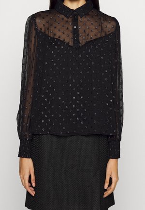 Mujer que lleva una blusa negra transparente con textura de lunares, cuello abotonado, mangas largas y una falda negra con textura.