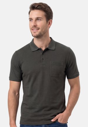 Mann trägt dunkelgraues Poloshirt mit kurzer Ärmel und Brusttasche, lächelt und schaut nach links, Hand in der Tasche der blauen Jeans, weißer Hintergrund.