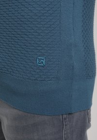 Maglione blu a trama con motivo a diamante, polsini a coste e una piccola etichetta con logo all'orlo. Materiale liscio e design aderente.