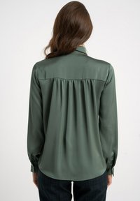 Bluzka z ciemnozielonego satyny z długim rękawem, plisowanym tyłem, mankietami na guziki i szpiczastym kołnierzem, zestawiona z spodnie w kratę.